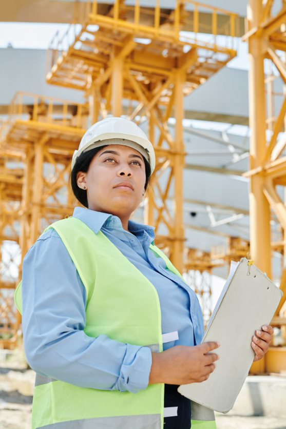 Single female construction worker wearing hard hat and high visibility jacket, looking into the distance and holding a clipboard
